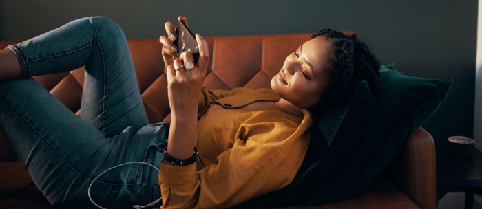 Woman laying on a brown couch in her living room, holding her Galaxy smartphone in landscape mode and she looks at the mobile gaming screen.