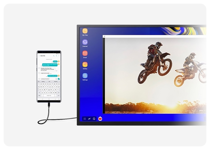 A Galaxy smartphone connected via USB cable to a desktop monitor to enable DeX. There is a photo on the monitor of two bikers mid-air. The smartphone shows a text message conversation on-screen