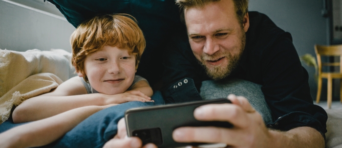 Father and son lay in bed on their bellies as the father holds a dark grey smartphone in landscape mode. Both the father and son smile as they gaze into the phone's display as if they're watching a show at home.