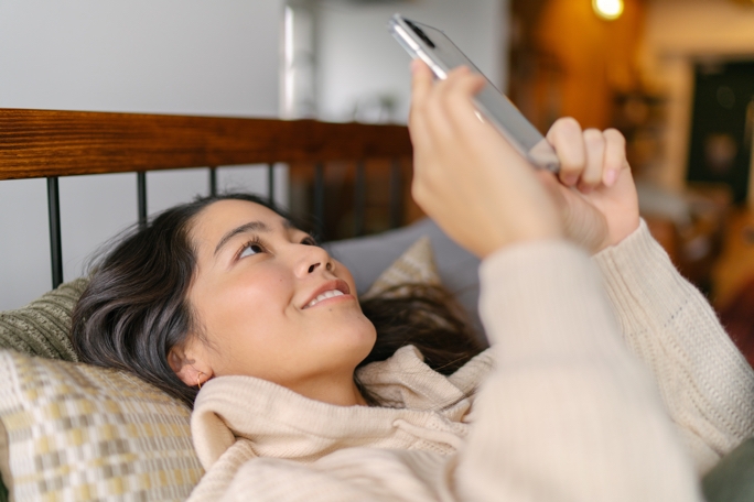 A lady laying on her bed in her home office, she is holding up a silver Galaxy S20 and smiling at the content on the screen