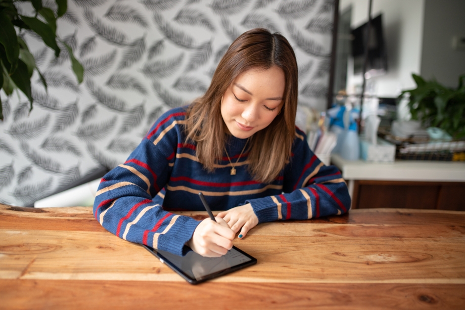 Amanda Rach Lee bullet journaling on her Galaxy Tab S6 Lite with a dark grey S Pen as she sits at a wooden desk. She's wearing a navy blue sweater with red and pink stripes.