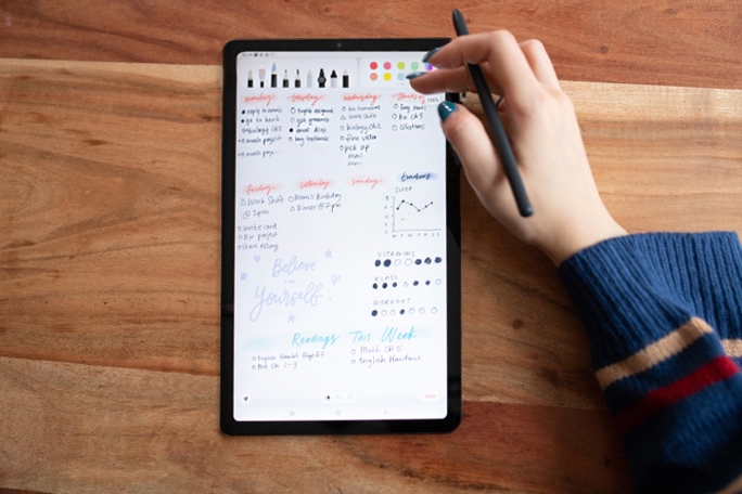 An overhead shot of a black Galaxy Tab S6 Lite that shows a weekly spread of plans, habit trackers, a motivational quote that says "Believe in Yourself" and a list of readings for the week on the Samsung Notes app. There is a range of pen/pencil options at the top left corner with a range of color options on the top right corner, which Amanda is navigating with her finger as she holds an S Pen.
