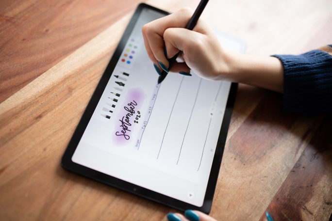 An overhead shot of a black Galaxy Tab S6 Lite angled slightly toward the left. Amanda's right hand is mid-design with a black S Pen. On-screen is a weekly spread for September 2020, created in Samsung Notes app.