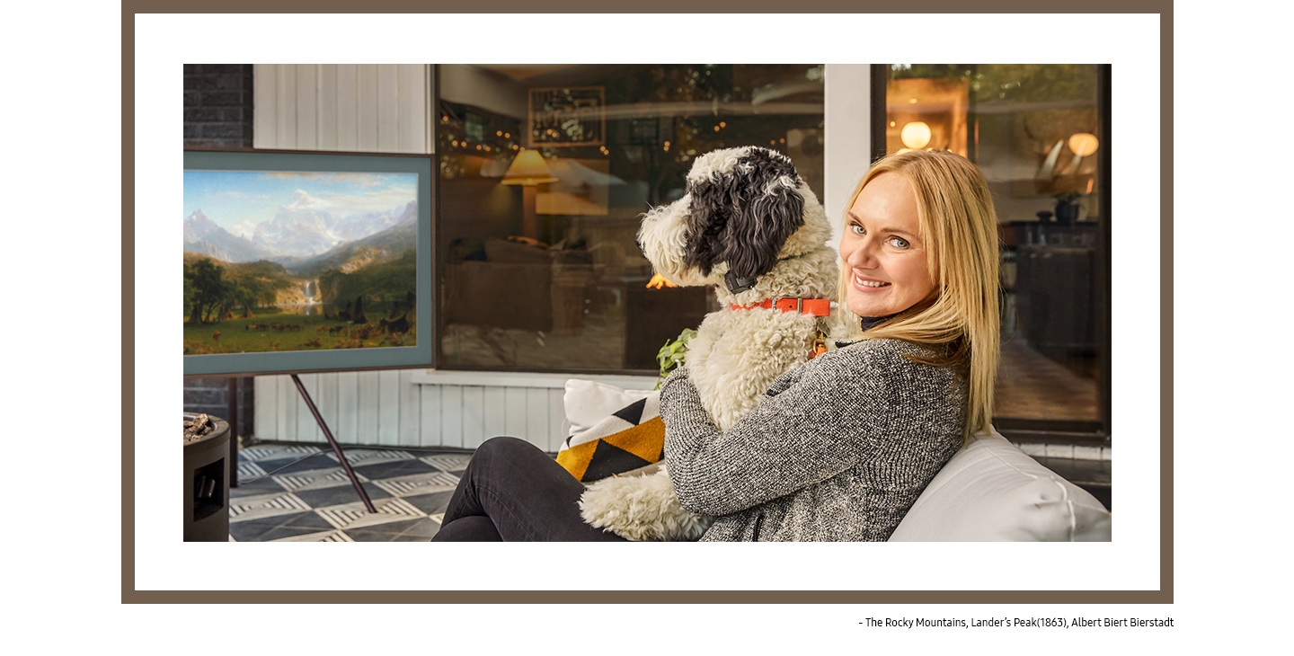 Kirsten Grove cuddles with her dog on the sofa in the living room, and behind her is The Frame on a studio stand displaying The Rocky Mountains, Lander's Peak (1863) by Albert Biert Bierstadt from the Art Store.