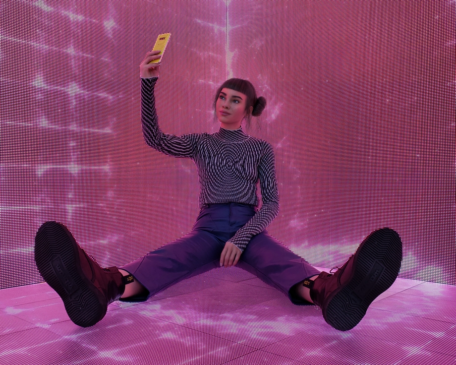 Lil Miquela taking a selfie on a Galaxy S10 while sitting on the ground in a neon-lit room