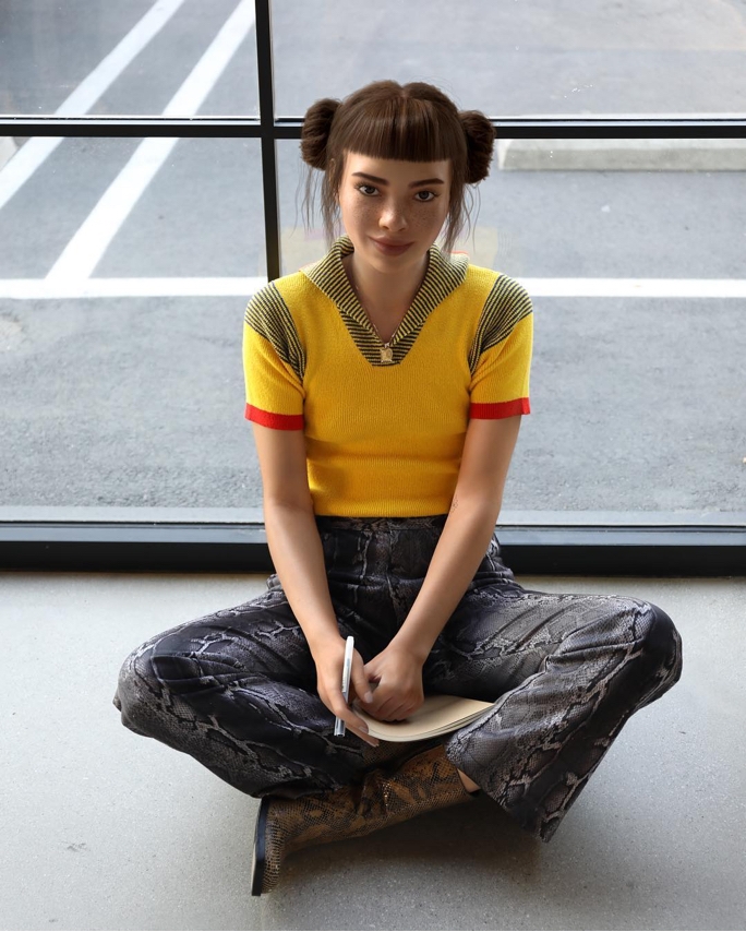 Lil Miquela smiling into the camera while sitting on the ground holding a pen and a notepad