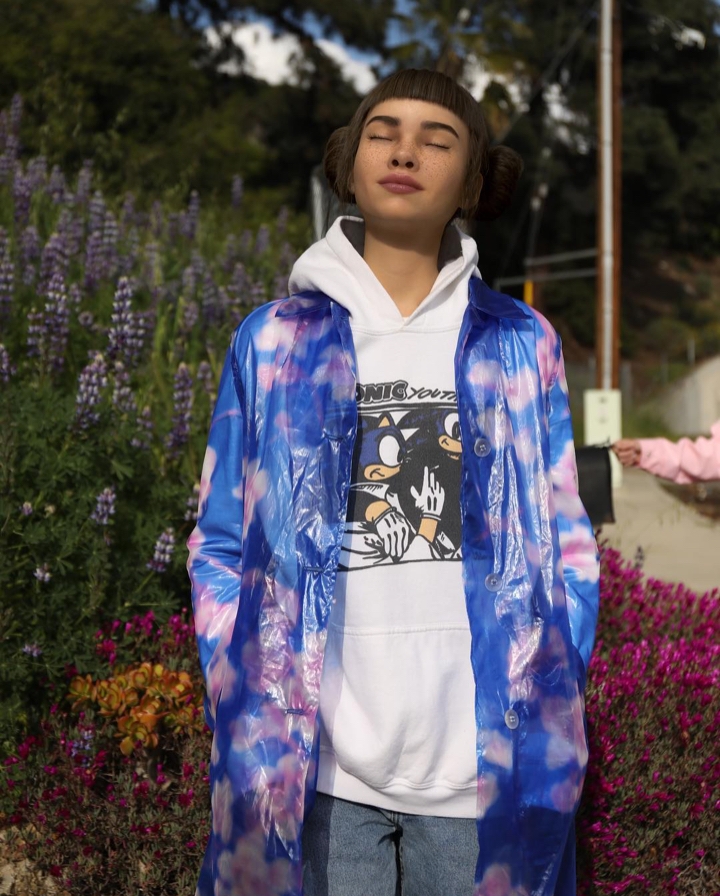 Lil Miquela standing outside among flowers, basking in the sun with her eyes closed