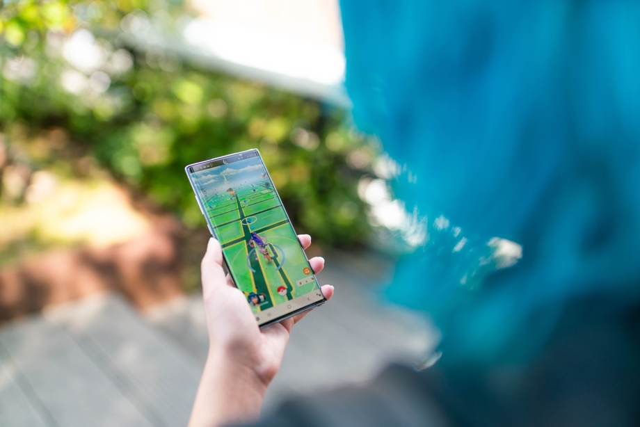 A lady standing outside and holding a phone with Pokemon GO visible on the screen. The image is shot from over her shoulder.