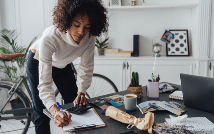 A lady in an apartment leaning over a desk and writing something on a piece of paper on a clipboard
