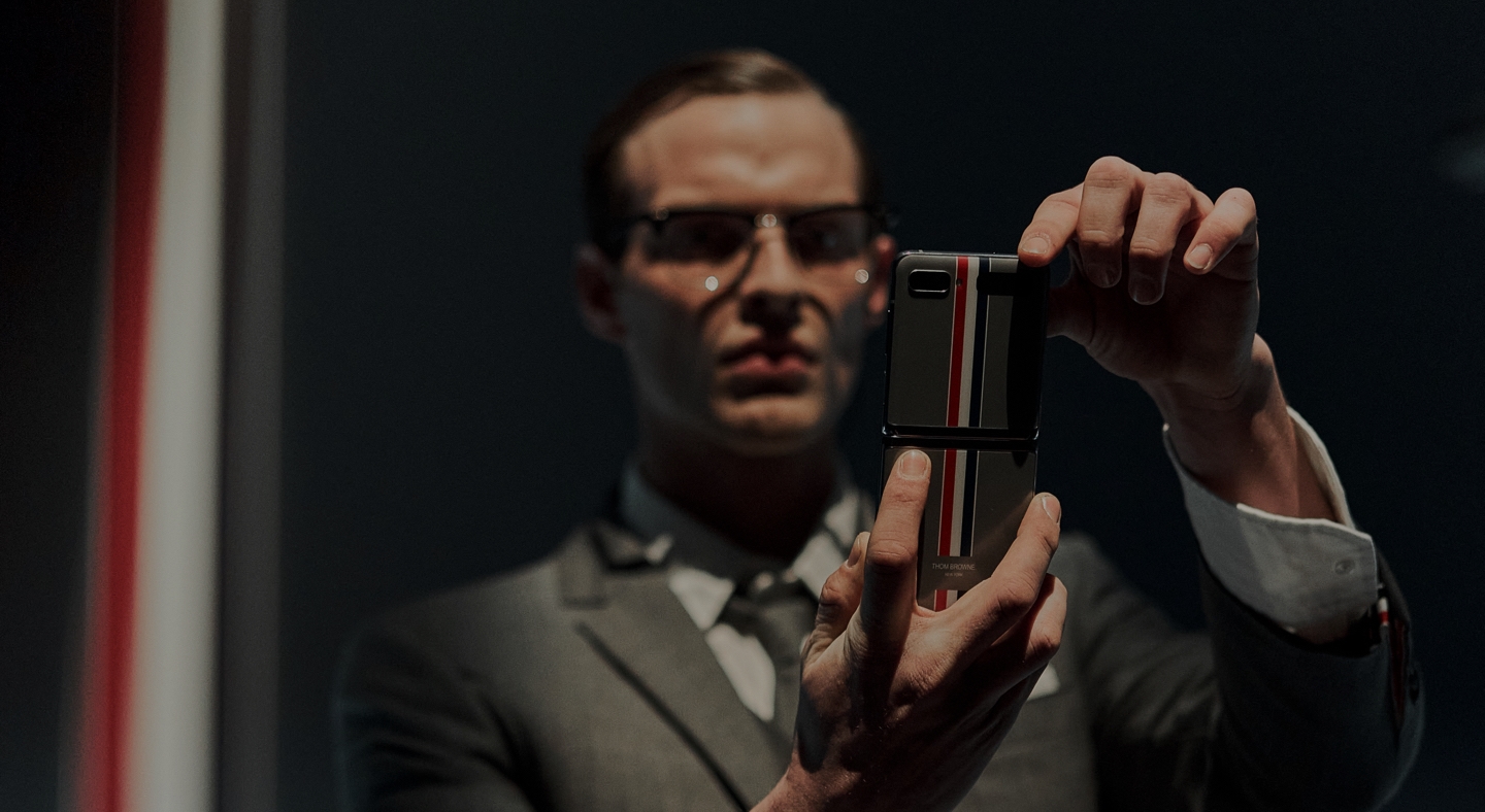 A male model is pictured standing and holding the Galaxy Z Flip Thom Browne Edition unfolded with both hands. He stares intently at the screen.
