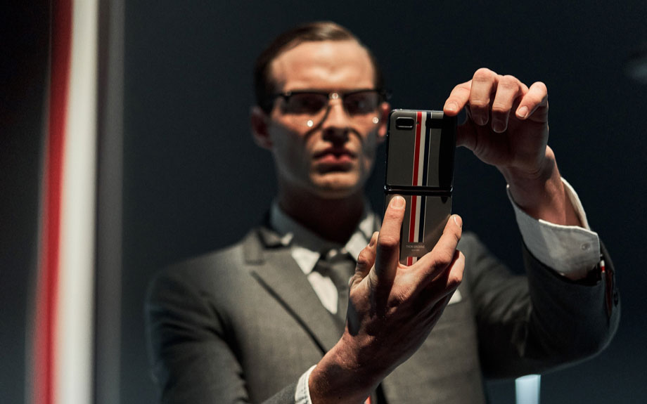 A male model is pictured standing and holding the Galaxy Z Flip Thom Browne Edition unfolded with both hands. He stares intently at the screen.