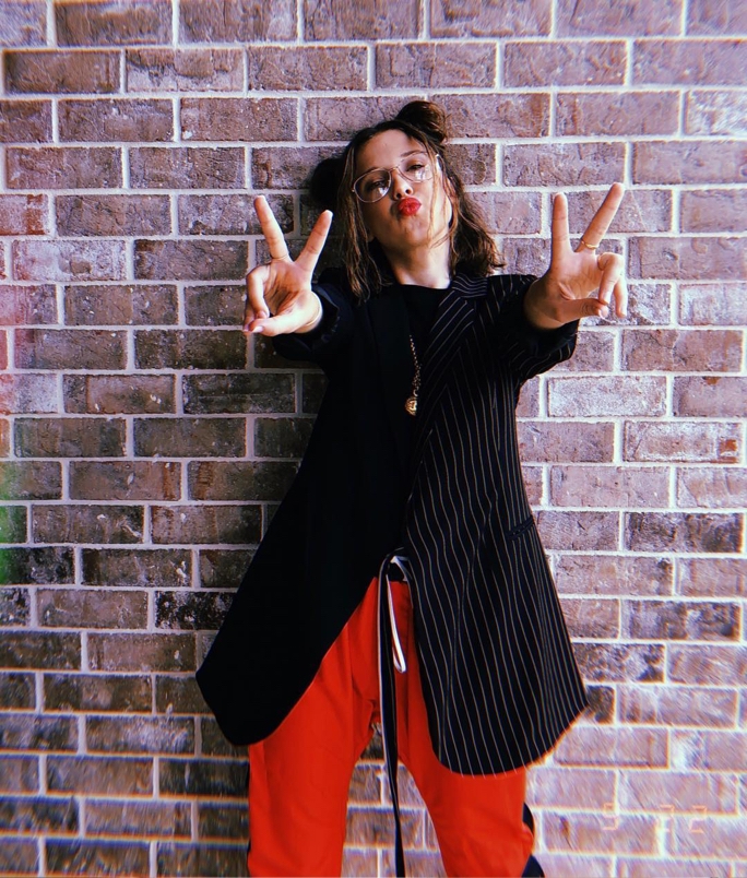 Millie Bobby Brown standing against a brick wall in a black pinstriped blazer and red pants, and forming two peace signs with her hands as she pouts her lips for the camera.