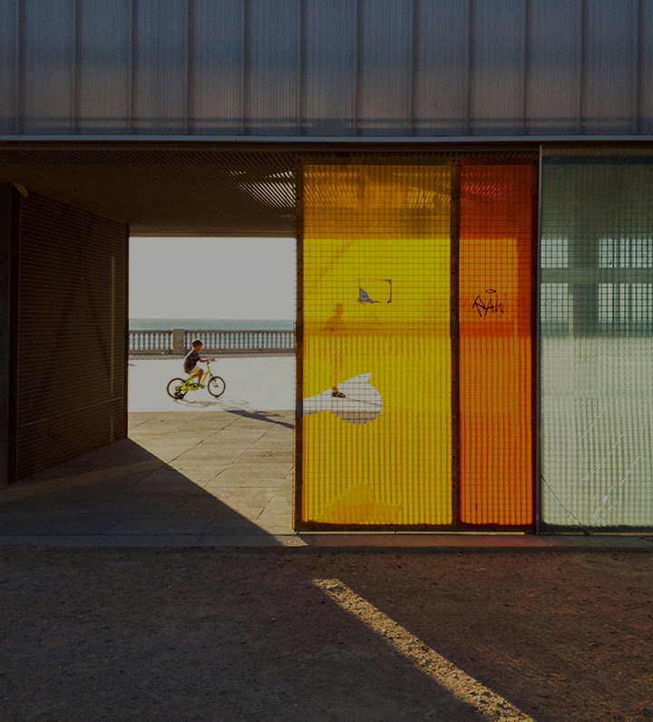 A little boy rides his bicycle on a boardwalk on a sunny day, the ocean as his backdrop. The view of him is captured inside an open building behind yellow, red and green window panels. A man in front of the boy looks toward the direction of the building.