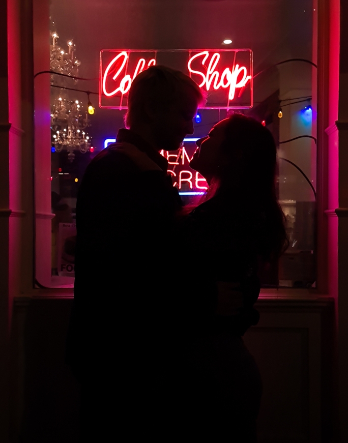 Silhouette photography of a couple standing in front of a neon coffee shop light, the scene is dark but you can make out their outline