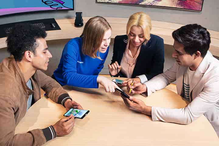Three people in their twenties at a table with a Samsung Experience store expert who is showing them how to use the S Pen with the Galaxy Note 9. Three people in their twenties at a table with a Samsung Experience store expert who is showing them how to use the S Pen with the Galaxy Note 9.