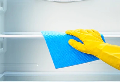 Hand wearing yellow glove using blue cloth to clean an empty refrigerator shelf Hand wearing yellow glove using blue cloth to clean an empty refrigerator shelf