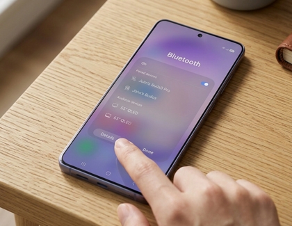 A hand tapping a Galaxy phone on a wooden table showing the Bluetooth settings screen with paired devices listed.