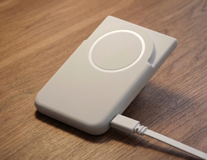 A gray Samsung battery pack lying on a wooden surface being charged by a white USB cable.