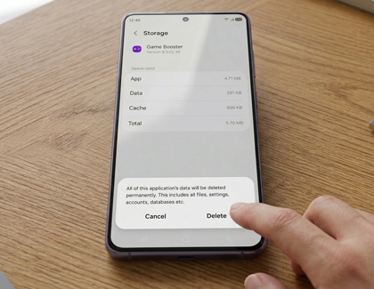 A hand tapping a Galaxy phone on a wooden table showing the Game Booster storage screen with a delete confirmation dialog.