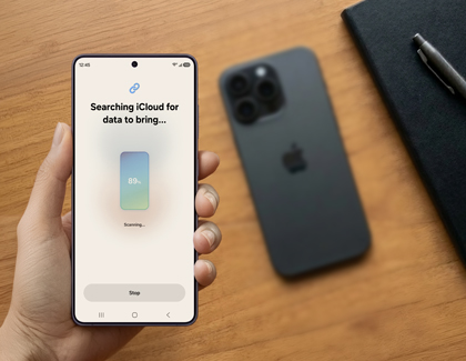 A hand holding a Galaxy S26 Ultra that is searching iCloud for data to bring next to a blurred black iPhone on a wooden desk.