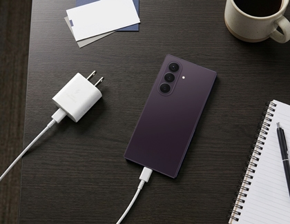 A dark purple Galaxy Z Fold8 phone plugged into a white wall charger on a dark wooden desk.