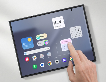 A hand touching an Ask Gemini widget on a Galaxy Z TriFold main screen with other widgets like Chrome Dino and a calendar floating above.
