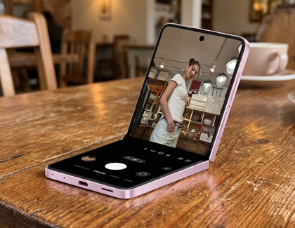 A pink Galaxy Z Flip8 standing partially folded on a wooden table in a cafe taking a photo of a woman.