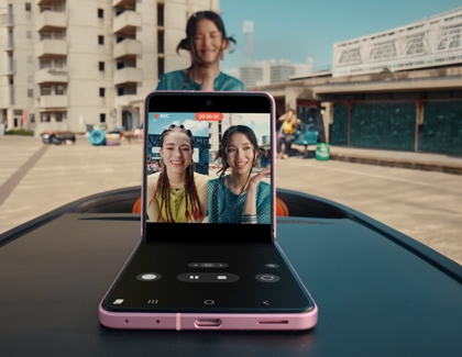 A pink Galaxy Z Flip8 standing partially folded on a dark surface outdoors recording two smiling women on the main screen.