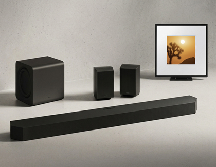 A black Samsung soundbar, subwoofer, and two rear speakers on a floor near a framed picture.