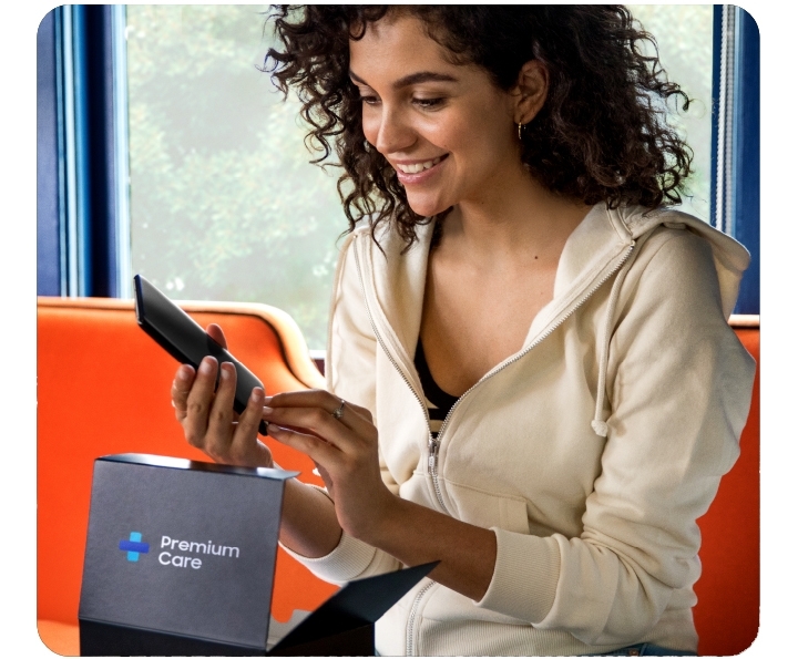 A smiling woman in a hoodie sitting on a coach with Premium Care box open on her lap. She is holding a Galaxy phone. A smiling woman in a hoodie sitting on a coach with Premium Care box open on her lap. She is holding a Galaxy phone.