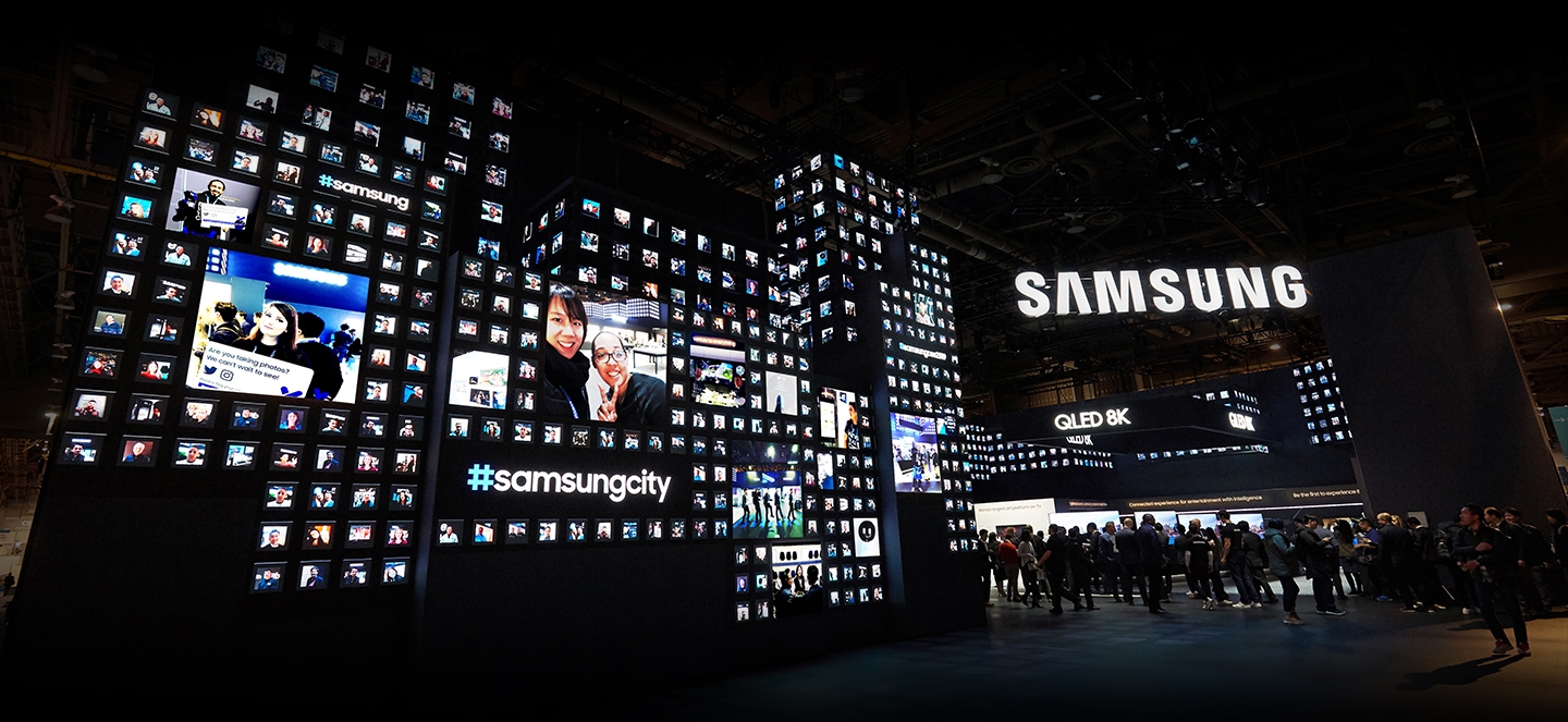 CES 2019 attendees enter the Samsung booth, where the massive front façade of the entrance is made up of multiple LED screens with one continuous display comprised of hundreds of small, tile-shaped photographs surrounding the hashtag 'Samsung city'.