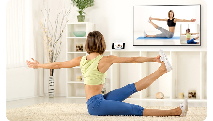 A woman, sitting down in her living room, is following a yoga pose showing on the TV in front of her. Next to the TV, her smartphone is recording her pose, which is being mirrored onto a small window on the lower righthand corner of her TV so that she can compare her pose to the instructional video playing.