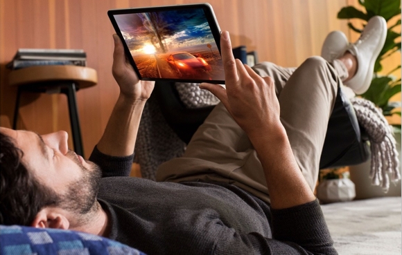 A man laying on the floor of his home, he is playing a driving game on his tablet. The image on the screen of his tablet shows a red car driving away from a tornado.