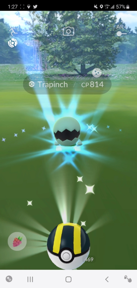 An in-game screen capture of a black and yellow striped pokeball in front of a pokemon hatching from an egg, surrounded by greenery.