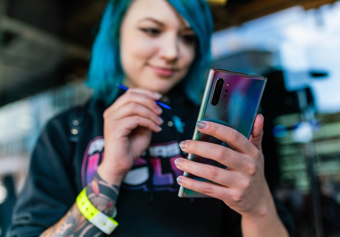 Holly holding an Aura Glow Galaxy Note10 in one hand with a blue S Pen in the other hand as she looks at the screen. The Galaxy Note10 is in focus while Holly is out of focus in the shot. 