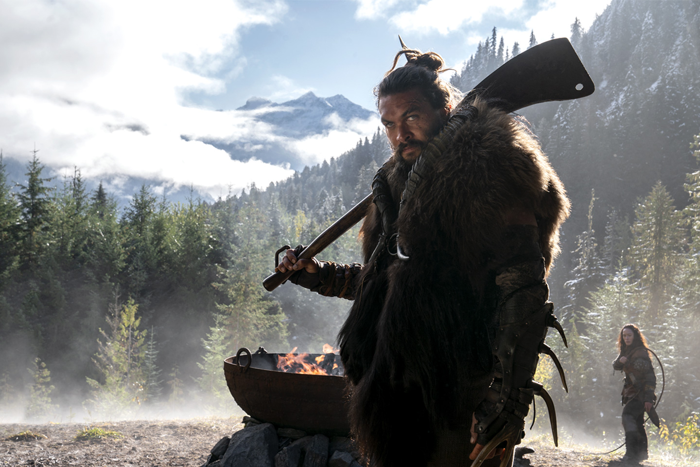 Scene from the Apple TV+ Originals show See that shows Jason Momoa's character looking off into the distance  while holding a weapon. He is in a forest and there is a man-made fire burning behind him.