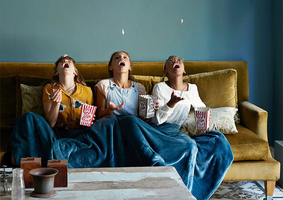 Three friends share a blanket on the couch while throwing popcorn up and catching it in their mouths. 