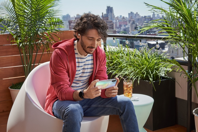 A guy sitting in a white egg chair on his balcony, holding a Galaxy Note10 phone in landscape mode. He's looking down at his phone while wearing a pair of white Galaxy Buds+ mobile gaming headphones. A skyline view can be seen in the distance. There are plants surrounding him.
