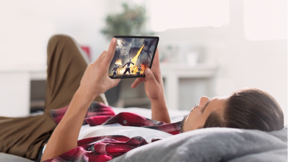 A man wearing a red flannel shirt and brown cargo pants is laying down in his bed holding up a black Galaxy Tab A 8.4 with both hands. On display is a capture of a game where three soldiers are in battle while a dragon is breathing fire toward them in a medieval setting. 
