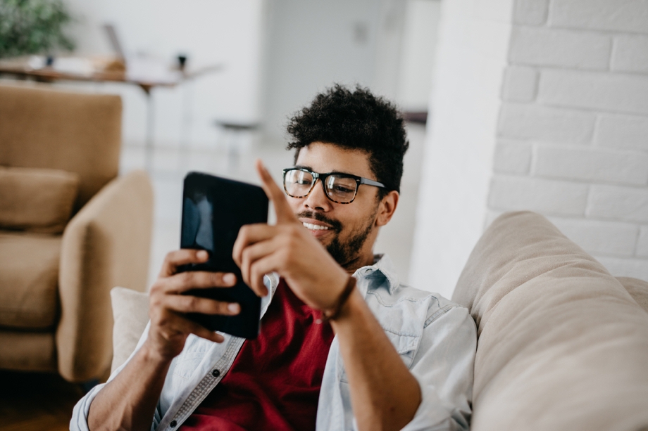 A man at home in his living room, laying on a beige couch. He's smiling while playing a game on his Galaxy tablet.