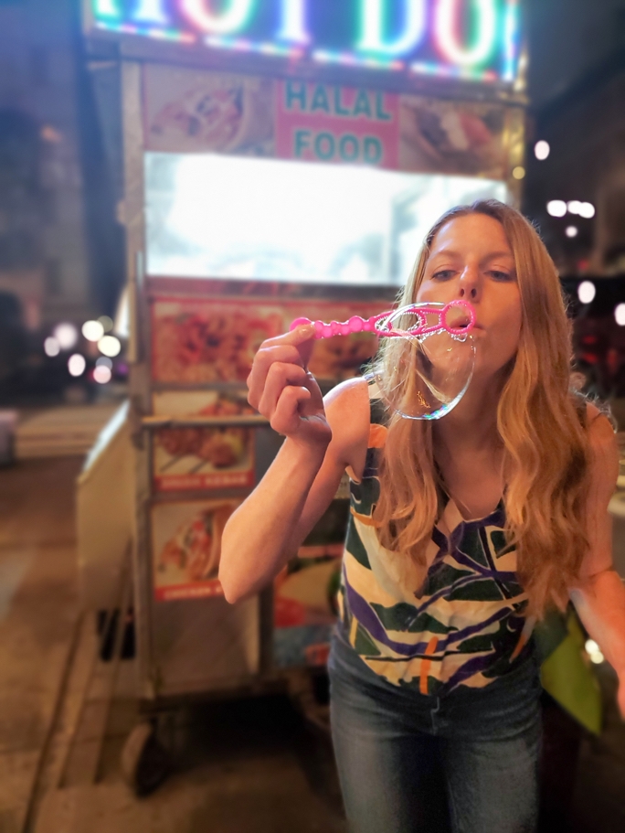 Bokeh night photography of a woman blowing a bubble while standing in front of a well-lit food cart 