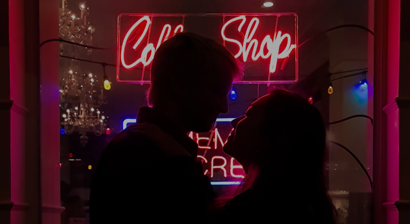 Silhouette photography of a  couple standing in front of a neon coffee shop light, the scene is dark but you can make out their outline