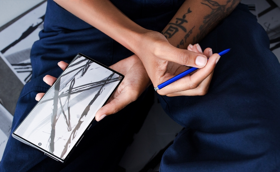 An overhead shot of a person with their arms crossed over their lap, they have a Galaxy Note10 in one hand and an S Pen in the other