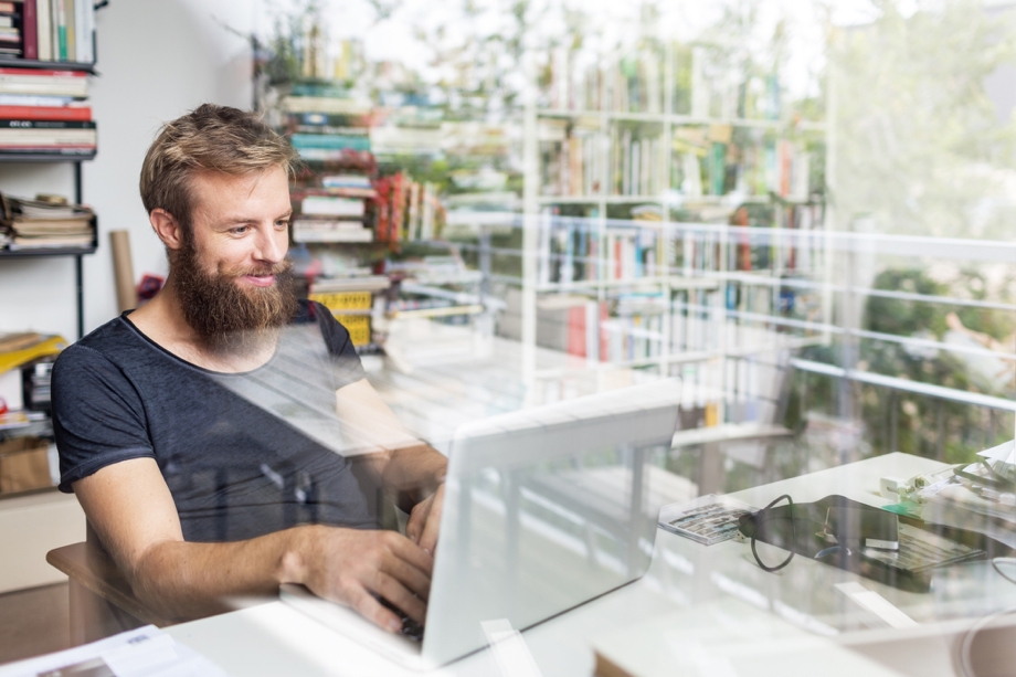 A double-exposure shot of a man sitting at a desk on his laptop, there is a book shelf in the background