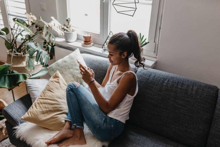 A lady sitting on a couch in a plant-filled room, she is on her phone