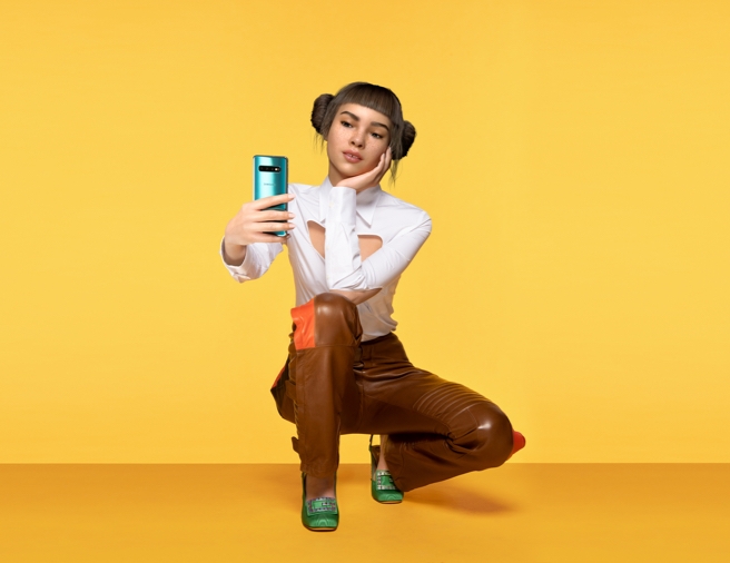Closeup shot of Lil Miquela against a pale yellow background as she squats down, holding a reflective blue Samsung Galaxy S10 smartphone