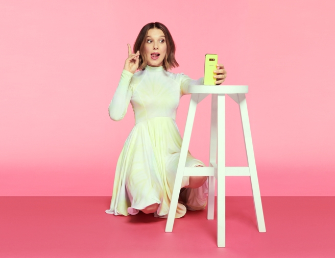 Millie Bobby Brown kneels while holding up a peace sign with her fingers as she takes a selfie with a neon yellow Samsung Galaxy A80 that's propped on a white stool