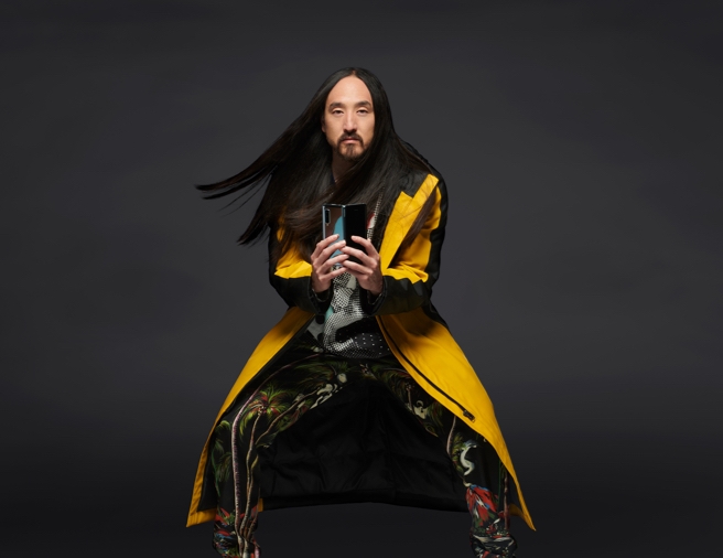 Steve Aoki standing with his knees slightly bent outward while sporting a yellow long coat as he holds up an unfolded Samsung Galaxy Fold smartphone 