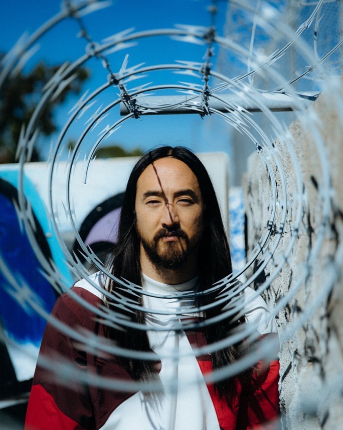 A headshot of Steve Aoki gazing seriously through the opening of a barbed wire fence in an urban yard setting