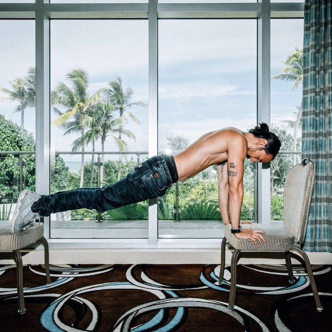 Steve Aoki uses two chairs to prop up his body for a fully-formed pushup in his hotel room with palm trees outside his window as backdrop for his resistance workouts